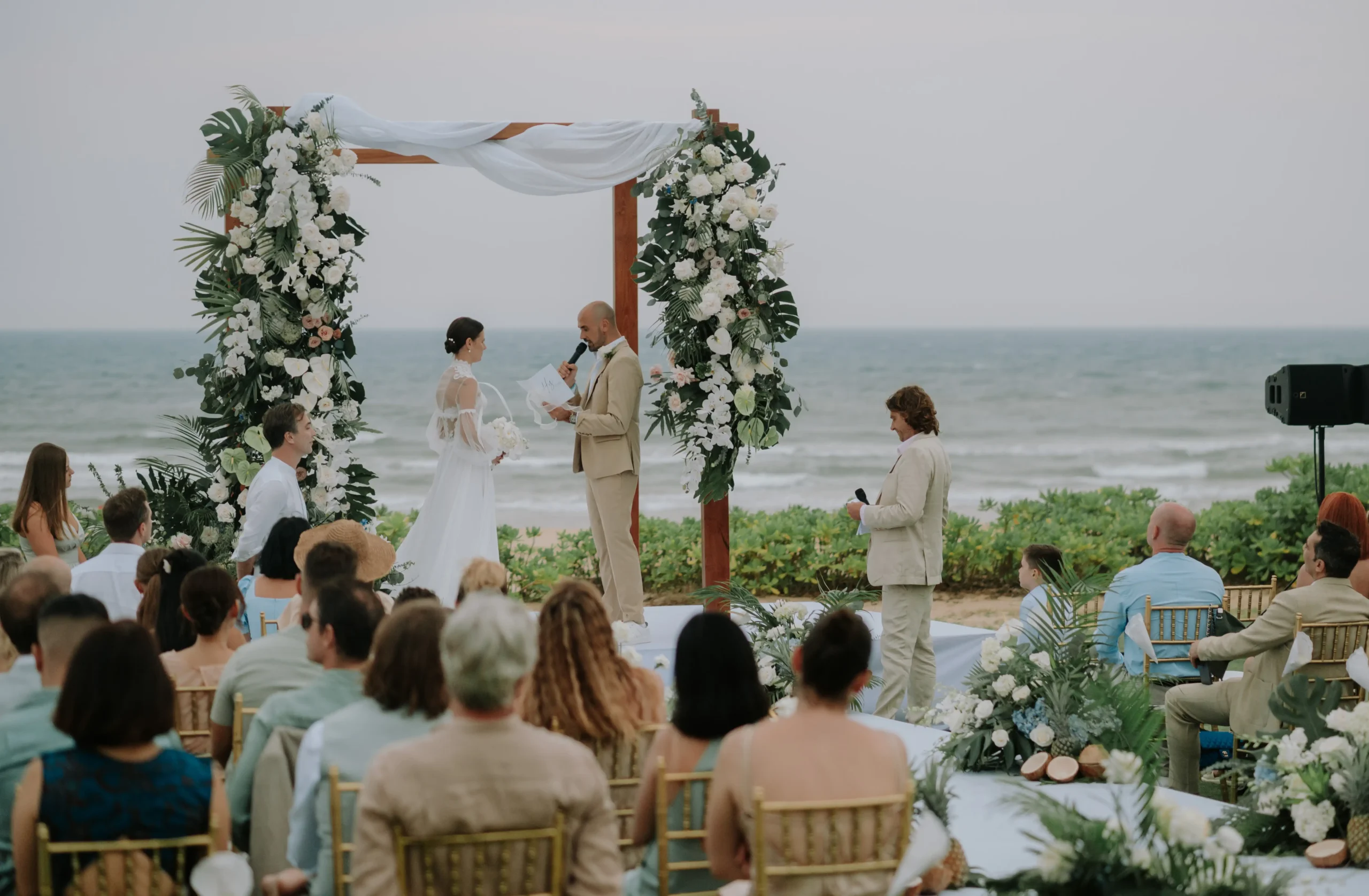 Guests watching a beach wedding ceremony under a floral arch at Bliss Hoi An