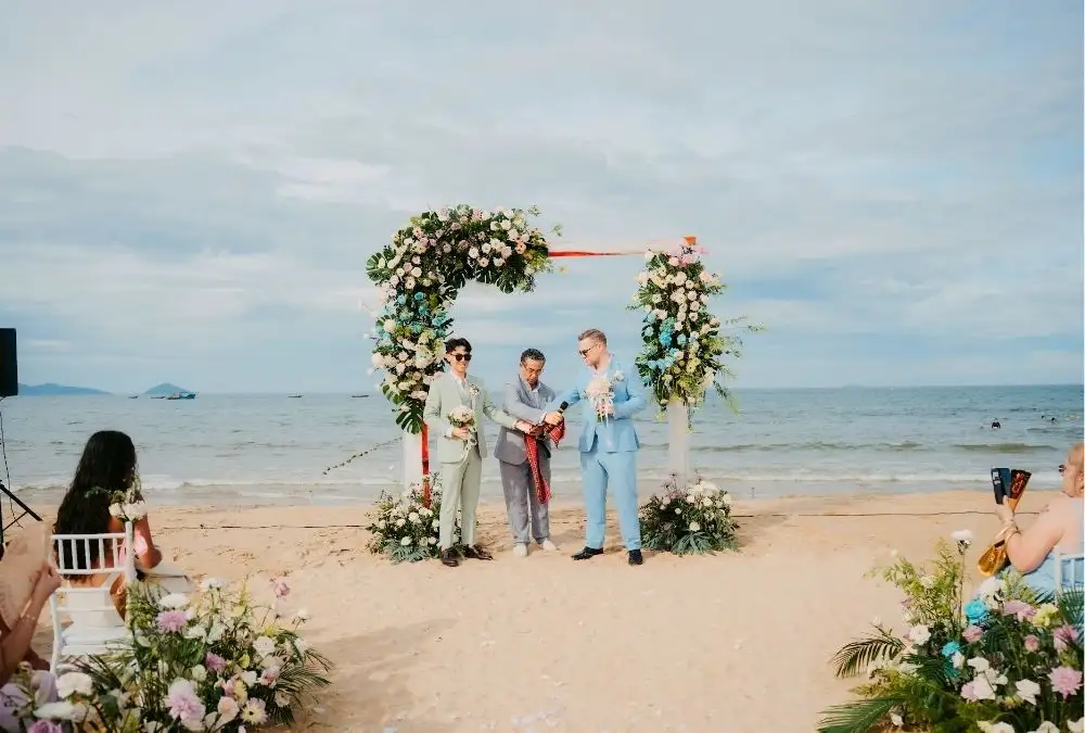 Daniel and Kieran stand before the floral arch with their officiant, the sea behind them